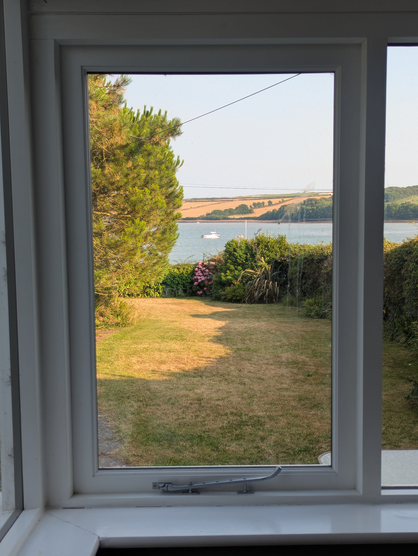 A window framed view from the house across the garden and pine tree to the bay beyond.